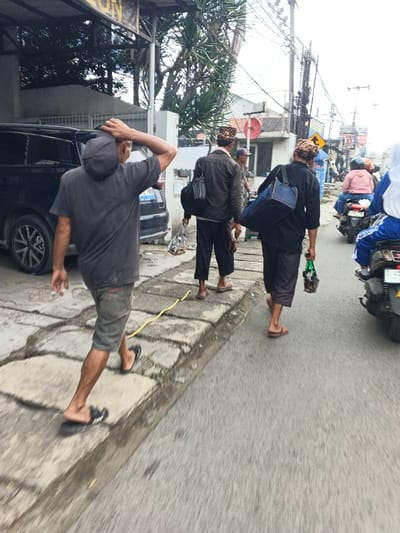 A street scene features people walking on a sidewalk and motorcycles traveling on the road. A man in a gray shirt, shorts, and flip-flops touches his head. Two men in traditional hats and dark clothing carry bags. Motorcycles with riders wearing helmets and a headscarf are on the right. Overhead wires are visible in the background. A black SUV is parked nearby.