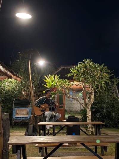 A young man with glasses is playing an acoustic guitar and singing into a microphone. He is sitting on a stool under an outdoor lamp in what appears to be a garden or patio area. The man is wearing a dark green jacket with white stripes on the sleeves, a black shirt, and light-colored pants. A black backpack is visible on the ground behind him, next to a blue and yellow cart. In the background, there is a tree with green leaves and red flowers, a building with a carved wooden facade, and dense foliage. The scene is illuminated by an overhead light and appears to be taking place at night. The location is identified as Campurejo, Indonesia.