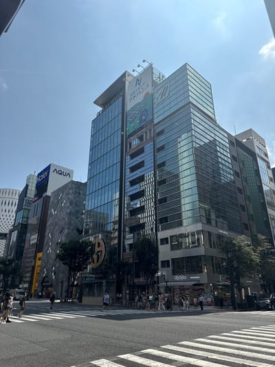 A sunny daylight scene depicts a busy urban intersection in Ginza, Chūō, Japan. The foreground is dominated by an asphalt road with a prominent white pedestrian crosswalk marked with parallel lines.

Numerous pedestrians are observed on both sidewalks and actively crossing the street. On the left, a smaller group of individuals walks on the sidewalk, with one person crossing the road from left to right. On the right, a larger group of pedestrians is gathered at the corner and in the crosswalk. A black SUV is parked or idling on the far right.

The background features multiple high-rise commercial buildings under a clear blue sky with sparse, wispy clouds. From left to right:
1.  A distinctive white building with a patterned, grid-like facade is partially visible on the far left.
2.  Adjacent to it, a building displays "Haier" and "AQUA" logos near its top.
3.  A dark gray building with a textured, diamond-patterned facade prominently features "BOTTEGA VENETA" vertically.
4.  A tall, modern glass-clad building bears "GIORGIO ARMANI" at its apex, with a large golden, stylized 'G' logo visible at street level.
5.  To the right of the Armani building, another tall structure features a large vertical billboard displaying "自然エネルギー アドバンス" (Natural Energy Advance) alongside an illustration of solar panels and a wind turbine, and a prominent "N" logo.
6.  At the street level of the central buildings, retail storefronts are visible, including "matsukiyo" (Matsumoto Kiyoshi drugstore) and a partially visible "KIEHL'S" sign.
7.  Further to the right, another multi-story building with glass and patterned facades is partially visible.
Trees with green foliage line the sidewalks.