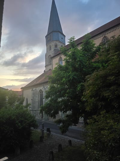 The image depicts the exterior of a large stone church with a tall, dark-colored spire that has a clock face. The church is surrounded by trees and greenery, with a cobblestone path leading towards it. The sky is overcast with some hints of sunlight peeking through the clouds. The scene appears to be in a town or city, with buildings and a street visible in the background. The overall impression is of an old, historic church in a somewhat overgrown setting. Based on visual cues such as architectural style and the presence of a clock tower, this is likely the St. Johannes Kirche in Münnerstadt, Germany.