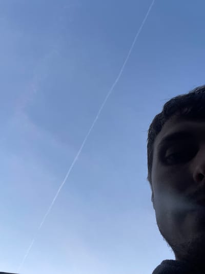 The image is a close-up, low-angle shot of a person's face and hair, partially obscured by shadow, against a clear blue sky with a thin white contrail. The person's right eye and a portion of their forehead and hair are visible on the right side of the frame. The contrail stretches diagonally across the sky from the lower left to the upper right. The image appears to have been taken outdoors, possibly in the city of Genk, Belgium, given the context. The lighting suggests it is daytime.