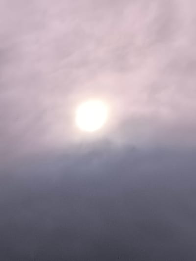 This media file is a vertical photograph taken outdoors, featuring a hazy sky with a bright, diffused sun prominently positioned slightly above the center. The sun appears as a luminous, almost white orb, its brightness creating a soft glow that permeates the surrounding atmosphere. The sky is filled with wispy, light-colored clouds that are tinged with a pale purple or pink hue, suggesting either a sunrise or sunset. The lower portion of the image is dominated by darker, denser clouds that create a gradient effect, transitioning from the lighter sky above to a deep, muted gray-purple at the bottom. There are no discernible people, objects, or man-made structures in the frame. The overall scene evokes a sense of atmospheric diffusion and muted light. The time of day is likely dawn or dusk, given the quality of light and cloud colors. The weather appears overcast or foggy, with the sun's rays softened by the atmospheric conditions. No text is visible in the image.