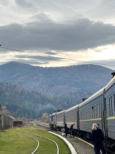 The scene depicts a train station in Mykulychyn, Ukraine, under an overcast sky with prominent grey clouds. In the background, forested mountains display a mix of green and autumnal brown foliage. A multi-car passenger train, primarily dark blue with light yellow accent stripes, is stationary on the tracks on the right side of the frame, alongside a low, curved concrete platform. Six individuals are visible on the platform: two in dark clothing stand in the right foreground with their backs to the camera, while four others are positioned closer to the train's entry points. One individual near the train exhibits a red garment detail. To the left of the tracks, a grassy expanse features multiple converging railway lines and several small, rustic buildings, including one with a corrugated curved roof. A pile of hay is visible near these structures. A faint plume of white smoke or steam emanates from the train's front portion on the left. Overhead electrical or communication lines traverse the upper sky.