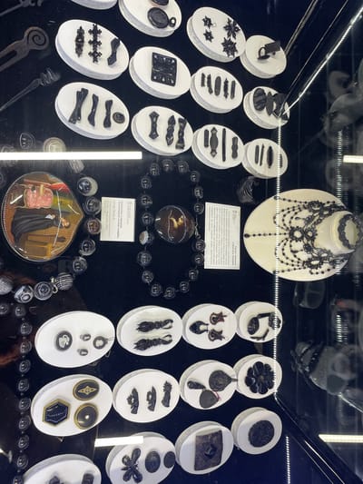 This is a top-down, high-angle shot of a display of antique jewelry and miniatures, likely at the Museum of Local Lore in Voskhod, Russia. The items are presented on white oval pedestals against a black background. Several items are displayed in glass cases, with vertical lighting strips visible on the right.

In the upper left quadrant, there are several rows of black, ornate brooches and hair accessories. To the right of these, a white oval pedestal displays a black, square-shaped item with a circular indentation.

In the middle of the image, a large, dark necklace with black beads and translucent, amber-colored pendant is displayed. To the left of this necklace, a heart-shaped miniature painting depicts a woman in a pink dress and a man in dark clothing, possibly within a domestic interior. A small information plaque is positioned between the miniature and the necklace.

To the right of the necklace, a white oval pedestal displays a more elaborate black beaded necklace and a bracelet. An information plaque is also present to the left of this display.

In the lower half of the image, various other black jewelry pieces, including brooches, earrings, and buttons, are displayed on white oval pedestals. Some of these pieces feature intricate designs and metallic accents. The overall impression is of a curated collection of historical decorative items.