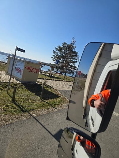 The image depicts a sunny daytime scene on Kiebitzweg, Kemberg, Germany, viewed from the interior of a white vehicle, primarily through its passenger-side rearview mirror assembly.
In the upper mirror, a reflection shows a person's right arm, clad in an orange high-visibility jacket, extending out of an open vehicle window with the thumb raised in a 'thumbs-up' gesture. A portion of the white vehicle's exterior is visible around the window. The lower mirror provides a similar but smaller reflection of the same arm and jacket.
The external scene, visible through and around the mirrors, features an asphalt surface in the immediate foreground, transitioning to a grassy area with sparse vegetation and bare ground patches. Cast shadows from unseen objects extend across the asphalt, indicating strong sunlight. To the left, a light-colored utility box, marked with red graffiti, stands beside a pole bearing a blue street sign displaying "KIEBITZ." Beyond this, a small wooden shelter with a dark roof is situated. A tall pine tree stands centrally in the midground, near a blue "P" parking sign. In the background, a large body of water, likely Bergwitzsee, stretches to the horizon under a clear, bright blue sky. A red circular stop sign is partially visible in the reflection of the upper mirror, positioned near the water's edge in the background.