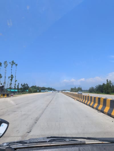 The image depicts a view from inside a vehicle driving on a highway. The road is concrete and appears to be a multi-lane highway with yellow and black striped barriers separating the lanes or road edges. The sky is clear blue with some scattered white clouds in the distance. On the left side of the road, there are several tall, slender trees, and beyond them, some buildings with green roofs are visible. On the right side of the road, more trees form a dense line. Several cars are visible on the highway, moving in the same direction as the viewer. The foreground shows the windshield and a portion of the dashboard of the vehicle, along with a side mirror on the left. The overall scene suggests a travel scene on a highway in a semi-urban or rural setting with vegetation.