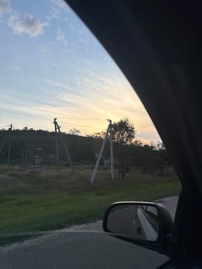 The image captures a rural scene, possibly on the outskirts of Sadaclia, Moldova, during what appears to be dusk. The viewpoint is from inside a vehicle, looking out the side window. The sky displays a gradient of colors, transitioning from a pale blue with scattered clouds near the top to a warm yellow and orange hue near the horizon, indicating the setting sun.

Dominating the middle ground are several power poles with visible electrical lines stretching across the frame. These poles are made of concrete and wood, some appearing old and weathered. They stand against a backdrop of a vegetated hillside, with trees and dense bushes obscuring much of the terrain. Some structures, possibly abandoned buildings or sheds, are partially visible amongst the foliage near the base of the hill.

In the foreground, to the right of the frame, a car's side-view mirror is prominent. The mirror reflects the road behind, showing a white line and the dark silhouette of the vehicle. The road itself is visible in the lower part of the image, appearing to be unpaved or made of gravel. A strip of green grass borders the road. The overall impression is one of a quiet, transitional moment in a rural landscape, marked by the presence of infrastructure and the fading light of day. No people are visible in the image.