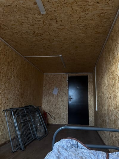 An interior shot shows a small, enclosed space with walls, floor, and ceiling covered in wood chipboard. A black rectangular storage unit is mounted high on the back wall. An extinguisher sits on the floor, near a stack of metal folding chairs. A white paper rests on the wall above the extinguisher. A clothes hanger extends from the right edge.