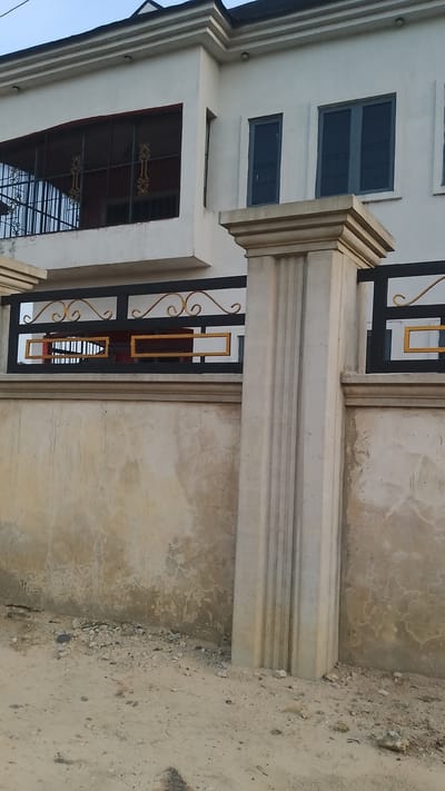 Exterior shot of a two-story white building in Asubi, Nigeria. The building features a balcony with a black metal railing on the second floor, as well as two visible windows. A concrete wall with a black metal fence and decorative gold accents runs along the perimeter of the property. The ground appears to be dirt or gravel. No people are visible.