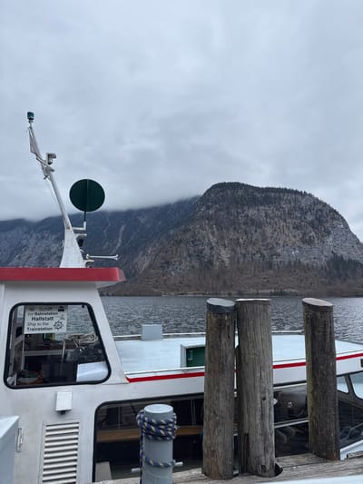 An elevated viewpoint captures a white passenger boat docked at a wooden pier on Lake Hallstatt, Austria, under an overcast sky. The boat, featuring a red accent stripe along its hull, occupies the left and central-lower portion of the frame. Its cabin window displays a sign reading "Schiff zur Bahnstation Hallstatt / Ship to the Trainstation." A white mast extends from the boat's foredeck, equipped with a green light, a small white flag, a circular green disc, and a metallic horn. On the wooden pier to the right, three weathered wooden mooring posts stand vertically; the leftmost post has a dark grey cap, the middle has a light grey cap, and the rightmost is uncapped. A grey metal bollard with a blue and white patterned rope coiled around it is positioned in the lower-left foreground on the pier. The dark, rippled surface of Lake Hallstatt stretches across the middle ground, with a steep, forested mountain range dominating the background. The upper slopes and peaks of the mountains are partially obscured by low-hanging grey clouds. No individuals are visible in the scene.