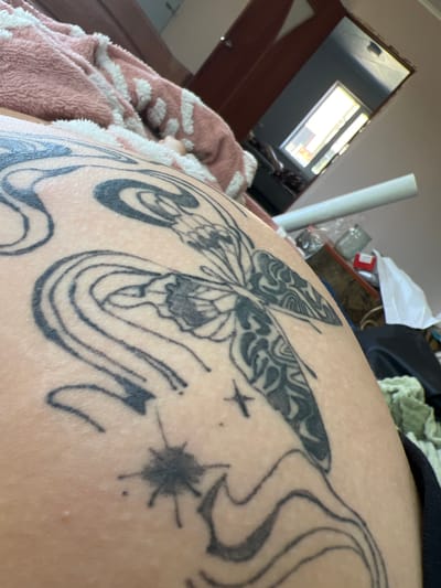 The image shows a close-up of a tattoo on a person's skin. The tattoo features a butterfly with intricate wing patterns, surrounded by swirling lines that resemble waves or smoke. There are also small star-like shapes and dots within the tattoo design. The skin appears to be lightly textured. In the background, a pink and white patterned blanket or robe is visible, along with glimpses of a room containing a mirror reflecting a window, and various objects such as a white tube, a glass jar, and folded fabric. The scene is lit by natural light coming from the window in the reflection. There is no indication of the city of Zudilovo, Russia.