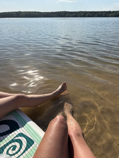 The photograph depicts a view from the perspective of someone sitting on a paddleboard with their legs dangling in the water. The location is likely a body of water near Yal'chik, Russia, though this cannot be definitively confirmed. The water is relatively clear, and the sun's reflection creates shimmering patterns on the surface and the sandy bottom can be seen through it. Two pairs of legs are visible, partially submerged in the water, extending from the paddleboard. One paddleboard is partially shown with a blue and white swirly pattern design on it. In the background, a line of trees appears to be along the shoreline. The overall impression is one of a relaxed, sunny day at the water.