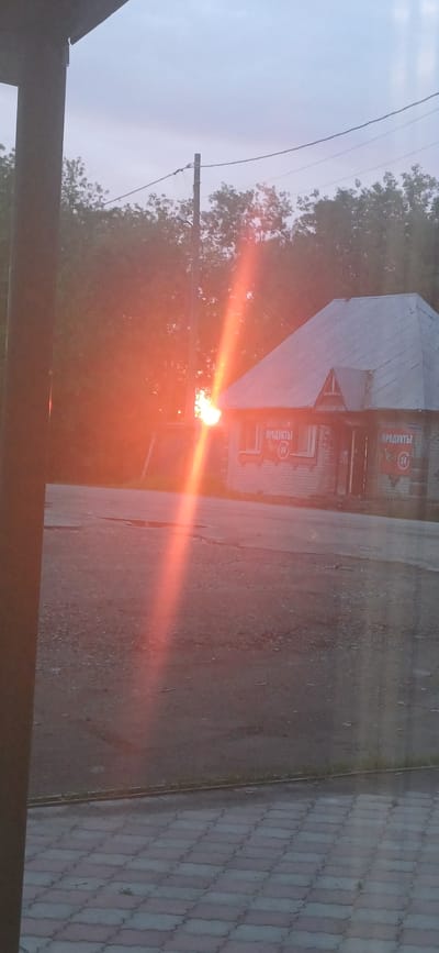 The image depicts a street scene in Zuevka, Russia, during what appears to be either sunrise or sunset. The sun, a bright orange orb, is positioned low in the sky and its rays create a prominent light flare diagonally across the frame, from the left foreground towards the center-right.
In the background, a small, single-story brick building with a gray metal roof is visible. The building has windows and signage that reads "ПРОДУКТЫ" (PRODUCTS) with a "24" symbol, indicating it's a 24-hour store. The building is situated on a paved area, likely a road or parking lot, which shows some wear and puddles. Trees line the background behind the building and extend to the horizon.
In the foreground, a section of a paved walkway with interlocking gray and reddish-brown tiles is visible at the bottom of the image. To the left, a dark, vertical post or structural element, possibly part of a porch or awning, obscures a portion of the scene. Power lines are strung across the upper part of the frame, supported by a utility pole. The sky is overcast with a pale, grayish-blue hue. No people are visible in the image.