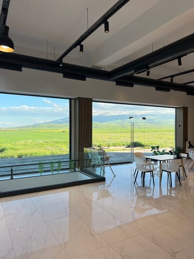 This is a high-angle, full shot of an indoor seating area with a large window overlooking a green landscape. The interior features a modern design with white tiled floors and exposed black ductwork and lighting fixtures on a white ceiling. In the foreground, a sleek table and several white chairs are arranged. The large window reveals a vast, rolling green field dotted with yellow wildflowers, under a partly cloudy blue sky. Distant mountains, some with snow-capped peaks, are visible on the horizon. A few small, green plants are displayed on a shelf-like structure along the window. The overall scene suggests a tranquil setting, possibly a cafe or restaurant, during daytime. The lighting implies it's a bright, clear day. The location is identified as Alagyaz, Armenia.