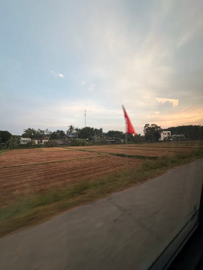 The image, when viewed in correct landscape orientation (rotated 90 degrees counter-clockwise), displays a rural or semi-rural landscape in Bình Chương, Vietnam, likely captured from a moving vehicle.
The sky occupies the upper half of the frame, presenting a soft, light blue color with subtle hints of warmer tones, suggesting either late afternoon or early morning light. A few faint, wispy clouds are visible, contributing to a generally clear sky.
In the foreground, along the bottom edge of the image, is a dark, greyish-brown surface, appearing to be a road or paved area. Adjacent to this, the mid-ground features extensive cultivated fields, predominantly brown and tan, indicating land that might be fallow, recently harvested, or prepared for planting. Some small patches of green are interspersed within these fields.
A line of diverse trees and dense foliage stretches across the middle of the landscape, behind the fields. Several small houses and structures are visible nestled among the trees and in the distance, including a notable multi-story white building. A tall, slender cellular or radio tower stands out against the sky in the background. A blurred, bright red, triangular-shaped object, possibly a flag or banner, appears in the mid-left portion of the image, suggesting movement or wind.
The scene depicts a calm, agricultural environment. No individuals are distinctly visible, and there is no discernible text in the image.