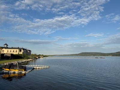 This is a wide-angle, eye-level, outdoor, daytime photograph of a lakefront property in the city of Kazashko, Bulgaria. The image captures a serene lake scene with a large, modern house on the left and a long pier extending into the water.
The house is a two-story structure with a beige facade, dark tiled roof, and multiple windows. It is surrounded by a manicured lawn and a rocky embankment leading down to the water. A single palm tree stands to the left of the house. In the foreground on the left, a yellow jet ski, covered with a black tarp, is docked on a floating pontoon. The pontoon is connected to a walkway that leads to the shore.
The lake itself is calm, with gentle ripples on the surface reflecting the blue sky and white clouds. A long, white pier stretches across the middle ground of the image, with several mooring posts visible. In the distance, rolling hills covered in greenery form the backdrop to the scene. The sky is a bright blue, dotted with scattered white clouds, suggesting a pleasant day. The overall impression is one of tranquility and luxurious waterfront living.