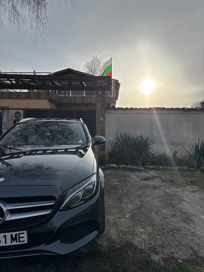 The image depicts the front left quarter of a dark-colored Mercedes-Benz sedan parked on an unpaved, dirt and gravel driveway. The car's front grille, headlights, and part of the license plate (ending in "1 ME") are visible. In the background, a two-story residential building, presumably in Bohot, Bulgaria, is partially visible. The building features a light-colored facade, a wooden pergola structure over a balcony on the upper level, and an air conditioning unit on the lower level. A Bulgarian national flag (white, green, red horizontal stripes) is flying from the roofline of the building, prominently displayed against a bright, hazy sky. The sun, a prominent bright spot, is positioned high in the right portion of the sky, casting lens flare artifacts. To the right of the car and in front of the building, a low, light-colored stucco wall with terracotta-style coping separates the driveway from a planted area containing spiky green shrubs. A stone pillar, likely part of a gate or fence, stands between the building and the wall, bearing the address number "24". Bare tree branches are visible against the sky in the upper left. No people are present in the scene.