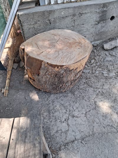 The image shows a weathered tree stump serving as a seat, located outdoors. The stump is rough, with visible bark and rings. Beside it, a bundle of dry grass, possibly a broom, lies on the ground. In the background, there is a concrete structure and some debris. A portion of a wooden plank is visible in the foreground. The scene appears to be in a rustic, outdoor setting, likely in the city of Kahramon, Uzbekistan, given the context provided. There are no people or discernable actions happening in the image.