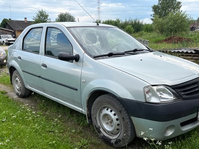 A light silver sedan is parked on a dirt and grass area, with a portion of another vehicle visible on the left. The car faces to the right, and its front is partially visible. The environment is rural, with a wooden building and a pile of firewood in the background. Trees and power lines are also present. The sky is overcast.