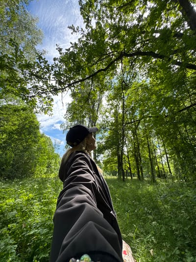 The image depicts a person wearing a black cap and a black jacket, seen from behind and to the side, looking up at the sky through a canopy of green trees. The person's hair is blonde. The environment is a lush, green forest with tall trees, thick undergrowth, and dappled sunlight filtering through the leaves. The sky is blue with scattered white clouds. There is no visible indication of this location being in Saint Petersburg, Russia.