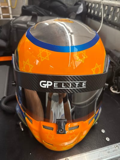 The image shows a close-up of a racing helmet painted orange, blue, and yellow, with the logo "GP ELITE" on a black carbon fiber visor. The helmet sits on a metal surface with a portion of a black bag visible in the background. The helmet appears to have been worn, as the visor is covered in a light layer of dust and dirt, and there are scratches on the surface of the helmet. The location of the photo is at the Circuit de Barcelona-Catalunya racetrack in the town of Montmeló, Spain, based on the provided coordinates.