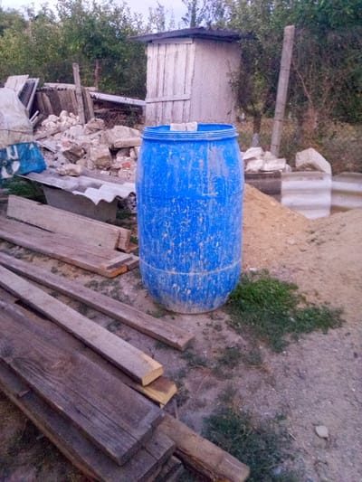 In Leușeni, Moldova, an outdoor scene depicts a construction or demolition site. In the foreground, several lengths of weathered wooden planks are stacked and scattered. Dominating the mid-ground is a large, bright blue plastic barrel, appearing somewhat worn and dirty, with a slightly open lid. Behind the barrel, a pile of rubble, including stones and broken concrete, is visible, alongside pieces of corrugated roofing material. Further back, a simple, light-colored shed or outbuilding with a dark roof stands, its walls possibly made of wood or metal siding. A chain-link fence runs along the right side of the image, partially obscured by overgrown vegetation. The ground is a mix of dirt, sand, and sparse grass. The overall impression is one of a disused or in-progress work area.