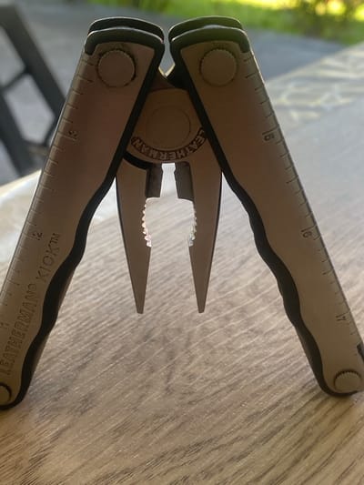 This is a close-up, angled shot of a Leatherman multi-tool. The tool is made of brushed metal with black accents and is resting on a light-colored, textured surface that appears to be a wooden table. The multi-tool is partially open, with the pliers extended. The jaws of the pliers are serrated.
Visible on the metal surfaces of the tool are engraved markings. On one handle, there is a ruler with markings up to "17". The numbers "15" and "16" are clearly visible. On the other handle, there is also a ruler, with numbers "3" and "12" visible. The brand name "LEATHERMAN" is engraved in a circular pattern on one of the handles, and "KICK™" is also visible.
The lighting suggests it is daytime, with a natural light source coming from the left, casting some shadows and creating highlights on the metal. The background is blurred, but shows hints of green, possibly foliage, suggesting an outdoor or near-window setting. There are no people visible in the image. The overall impression is one of functionality and practicality.