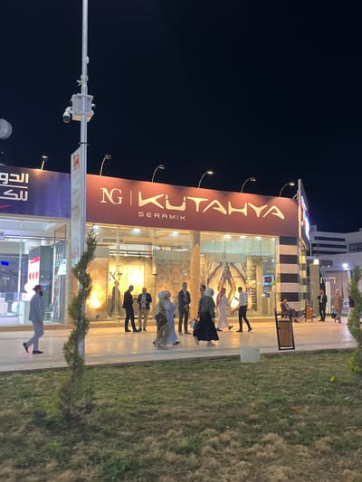 The image depicts a nighttime street scene in Al-Ghizlaniya, Syria, featuring a commercial building with a prominent sign reading "NG KUTAHYA SERAMIK". The building has large glass windows showcasing interior displays of what appear to be tiles or decorative materials. Several people are visible walking in front of and entering the establishment. One individual in a light blue suit is walking towards the left, while others, including individuals in traditional Middle Eastern attire and women in hijabs, are walking and interacting near the entrance. Streetlights illuminate the area, and a tall pole with security cameras is visible on the left side of the frame. The ground in front of the building is paved, with a grassy area to the foreground.