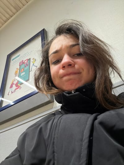 A person with brown hair and a grey jacket is looking down at the camera. They have a slight smile and their eyes are open. Behind them, there is a blue-framed poster on the wall, and to the right, a white wall with a metal strip. The image is rotated to the left.