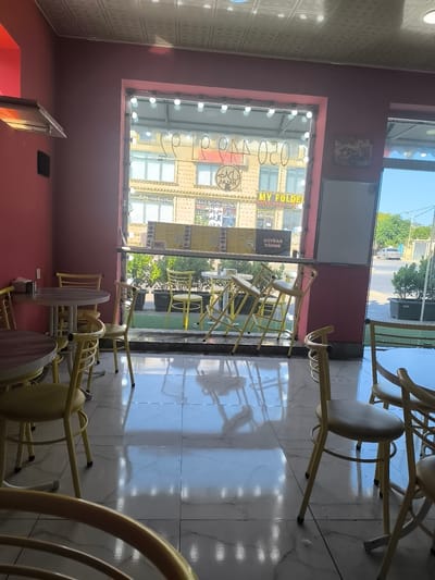 The image shows the interior of a fast-food restaurant with a window looking out onto a street. The interior walls are painted pink, and the floor is tiled with a marble pattern. There are several tables and chairs throughout the restaurant. Outside the window, there are buildings, trees, and a street with a car.
Specifically, the scene appears to be at a fast-food restaurant in Bina, Azerbaijan, indicated by the Azerbaijani text on the signage visible through the window. The restaurant has a modern, minimalist design with primarily pink walls and simple, yellow metal chairs and tables. The window provides a clear view of the street, showing a typical urban environment with other buildings, vegetation, and a vehicle. The overall impression is of a clean, functional establishment.