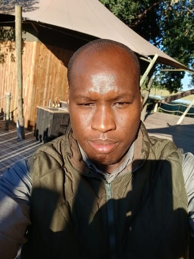 A close-up, tilted image of a man with dark skin, wearing a quilted green vest over a light gray shirt. He is outdoors; the background shows a wooden building or structure, and a light-colored canvas or shade sail, with green trees visible beyond. Sunlight casts shadows across his face and clothing. The exact location within Nwanaeanoka, Botswana, cannot be determined with the given information.