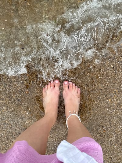 A first-person perspective shot from above shows a person's bare feet standing on a pebbled shore where shallow, clear water is lapping. The person is wearing a pink knitted garment on their legs and a white accessory that wraps around their lower leg. A pearl anklet is visible on the right ankle. The scene appears to be a beach, possibly in Derbent, Russia, given the context. The water is creating frothy waves as it washes over the pebbles.