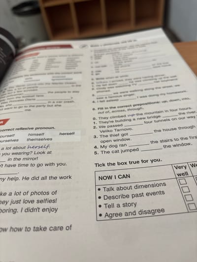 The image displays an open book on a desk, featuring various English language exercises. The book has a clear layout with sections for grammar, vocabulary, and comprehension. One section focuses on reflexive pronouns, with options like "yourself," "himself," and "herself" presented. Another exercise involves filling in prepositions such as "up," "down," and "into." A "NOW I CAN" section contains checkboxes next to phrases like "Talk about dimensions" and "Describe past events," suggesting a self-assessment activity. The environment appears to be indoors, with a wooden desk surface and a blurred background suggesting shelving units. There are no visible people or specific geographic landmarks, including any related to Dimovo, Bulgaria.