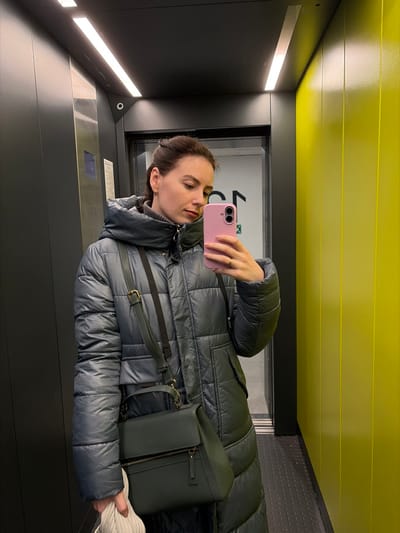 The scene depicts an interior view of an elevator in Ковалёво, Russia. A female subject is shown taking a mirror selfie, holding a pink-cased smartphone in her right hand, which displays a ring on the fourth finger. Her left hand grasps a folded light-colored garment. She wears a long, dark grey puffer coat over a grey turtleneck, and her dark brown hair is partially tied back. A dark green shoulder bag with a visible front zipper pocket is strapped over her left shoulder. The elevator features dark grey vertical paneling on the left and rear walls, with bright yellow vertical panels on the right. Overhead, two linear LED light fixtures illuminate the space. A small, circular security camera is mounted on the rear wall, above the dark elevator door frame. Through the partially open elevator door in the background reflection, a lighter-colored hallway is visible, featuring a large dark graphic symbol and a green illuminated exit sign on a white wall. A control panel with illuminated blue buttons is partially visible on the left wall.