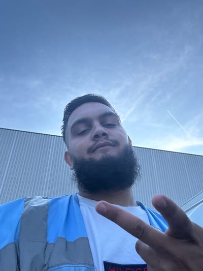 A low-angle, waist-up, selfie-style photograph features a young man with a full beard and dark hair, looking directly at the camera. He is wearing a white t-shirt and a blue and grey utility vest. His left hand is raised, with two fingers extended in a peace sign, while his right hand is blurred and out of focus. Behind him, a corrugated metal building forms the backdrop. The sky above is a pale blue with scattered white clouds and a faint contrail from an airplane. The location is the city of Boom, Belgium.