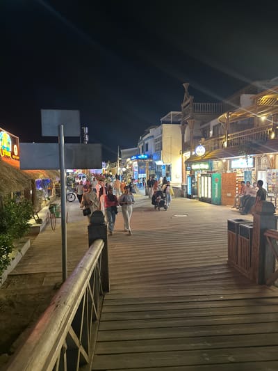 The image depicts a street scene in Dahab, Egypt, at night. A wooden boardwalk runs along the right side of the image, lined with shops and restaurants on the left. People are strolling along the boardwalk, some walking alone and others in groups. A large Heineken sign is visible among the shopfronts. Streetlights illuminate the scene. Some trees and vegetation are in the foreground.