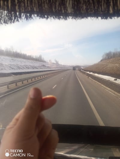 The image, taken from inside a vehicle, shows a highway stretching into the distance with snow-covered embankments on either side. A hand is visible in the foreground making a "finger heart" gesture. A truck can be seen further down the highway. The text "TECNO CAMON 19 Neo" is present in the bottom left corner. Based on the environment and the text, the image likely originates from the Chishmabash area in Russia, although without further context (time and date) it is not possible to determine the specific location.