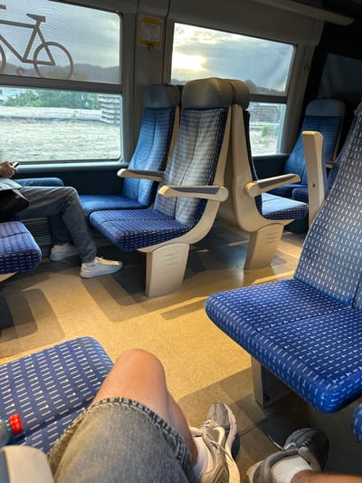 The image depicts the interior of a train carriage, likely in Alès, France, given the context. The seats are upholstered in a blue fabric with a white geometric pattern. The view from the window shows a landscape with a cloudy sky and some distant trees. In the foreground, a person's legs are visible, clad in denim jeans and white sneakers, resting on the floor of the train. The person is holding a smartphone. Several empty seats are arranged in rows, some with their backs to the viewer. The lighting suggests it is daytime, possibly late afternoon or early evening, as the sun is low in the sky. The overall scene is one of transit and quiet observation, typical of a train journey.