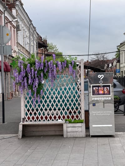 A photo booth stands on a paved street in front of a weathered building. The building's facade is brick, with layers of window frames and a sloping roof. A street sign is partially visible on the left. In front of the photo booth is a wooden structure, possibly a bench with a lattice back. The lattice is adorned with vibrant purple wisteria. A car is parked behind the photo booth. The sky is overcast. The photo booth is labeled "#ФОТОЛАВ" and includes Russian text mentioning Vladikavkaz. Based on the metadata, the location is identified as Akhieli, Georgia.