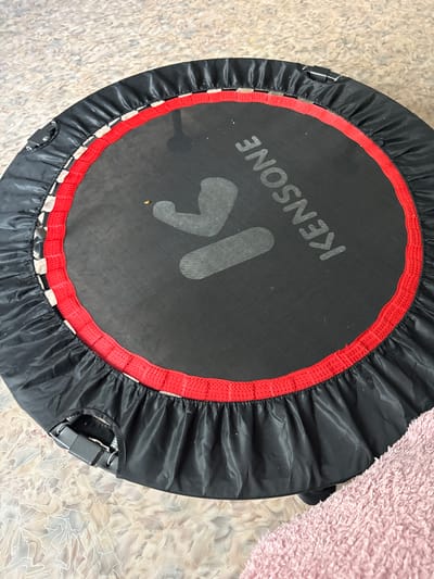 A close-up, overhead shot shows a black and red mini-trampoline on a textured floor. The trampoline has a black safety mat with a red edge, and a black skirt that covers the springs. The brand name "KENSONE" is visible in white on the trampoline mat. There are two grey, stylized figures printed on the mat, resembling a person in a jumping or stretching pose. A small yellow speck is also visible on the mat. One corner of the trampoline has a black strap with a buckle mechanism attached to the frame. A pink fuzzy blanket is visible in the bottom right corner of the image, partially obscuring the trampoline. The background floor appears to be a light-colored, patterned carpet or linoleum. The image is taken indoors, with no indication of a specific location in Айет, Kazakhstan.