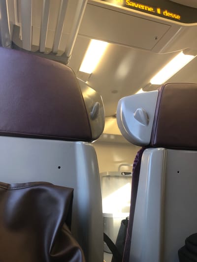 This image depicts the interior of a train carriage, located in Hochfelden, France. The foreground is dominated by the backs of two passenger seats. The left seat, partially visible, is upholstered in a dark reddish-brown material, featuring a light beige plastic shell. A dark brown, folded, soft material object, possibly a bag, rests at the lower left. The right seat displays an identical dark reddish-brown upholstery and light beige plastic shell; its top portion of the shell incorporates a small, integrated, ear-like ergonomic grip or design element.

Between the two foreground seats, a gap reveals a lighter grey interior structure, possibly a partition or a small utility cabinet, with a silver-colored horizontal handle visible near its top. A strong, diagonal beam of natural light illuminates this structure. A black fabric strap, likely from a bag, is visible at the bottom right, positioned against the base of the right seat.

The ceiling is off-white, featuring multiple rectangular, recessed LED or fluorescent light fixtures providing artificial illumination. Overhead luggage racks with a ribbed underside are visible above the left seat. An amber-colored LED display panel on the ceiling shows partial French text: "Saverne. Il desse". No individuals, specific actions, or interactions are visible within the scene. The environment is that of a clean, well-lit passenger train interior.
