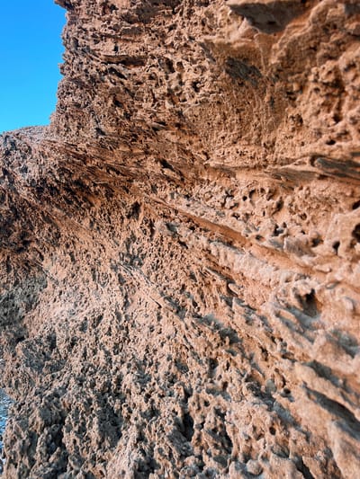 The media file is a close-up, low-angle shot looking up at the underside of a rocky overhang or archway. The rock surface is rough, porous, and textured, with many small holes and crevices, suggesting it might be weathered volcanic rock or a similar geological formation. The color of the rock is predominantly a warm, earthy brown with lighter tan and darker grey-brown shadows, indicating it's likely illuminated by natural light. The lighting suggests it is daytime, possibly late afternoon given the warm tones and shadows.
A portion of a clear, bright blue sky is visible through the opening of the arch, providing a stark contrast to the textured rock. The image is a detail shot, focusing entirely on the geological formation and the sky. There are no people, man-made objects, or specific activity depicted. The setting appears to be a natural outdoor environment, possibly a coastal area or a cave entrance, given the weathered rock and open sky. The lack of any recognizable landmarks or human presence implies a focus on the natural textures and forms of the landscape. No text is visible in the media file.