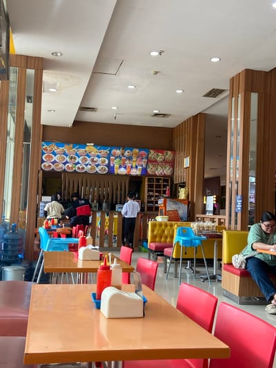 The image depicts the interior of a casual dining establishment in Kudus, Indonesia, likely a fast-food restaurant or a food court stall. The scene is brightly lit and features a variety of seating arrangements, including orange tables with red chairs, and yellow booth seating. A large menu board displaying various food items and drinks is prominently displayed on the back wall, above a counter area where employees are visible. Several employees, dressed in uniforms or casual attire, are attending to customers or preparing food behind the counter. In the foreground, tables are set with condiments like ketchup and chili sauce dispensers, and tissue holders. Water cooler bottles are visible on the left side of the image, suggesting a self-service water station. The overall ambiance is one of a busy, everyday eatery.