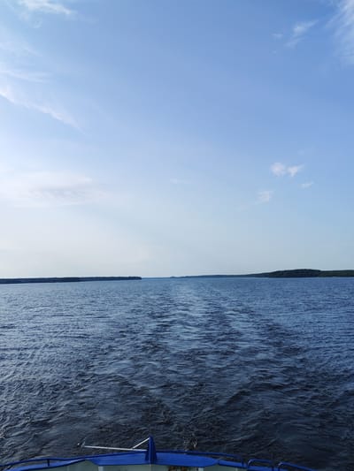 The image depicts a wide expanse of deep blue water under a clear sky with scattered wispy clouds. The water's surface is disturbed by ripples and a visible wake trails behind the viewer, suggesting movement. In the distance, a tree-lined shore stretches across the horizon, forming a dark, solid line against the lighter sky. The foreground shows the edge of a boat, with blue railings and white structures visible. The scene appears to be set on a body of water, possibly a lake or river, near Bessolovo, Russia. The overall impression is one of open space and tranquil travel on the water.
