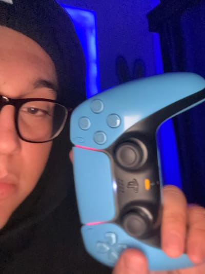 The image depicts an indoor scene focused on an individual holding a video game controller. On the left side, a person is partially visible from the nose upward, wearing dark-rimmed glasses and a dark hooded garment, looking directly towards the viewer. The person's right hand is holding a light blue PlayStation 5 DualSense controller in the foreground. The controller displays four white face buttons, two analog sticks, a PlayStation logo, an illuminated orange indicator light, and red light strips along its side edges. The background is dark, illuminated by a prominent blue light source, likely LED lighting, from the right. A faint, dark silhouette, resembling two upright, rounded forms, is visible against the blue background light in the upper right. The specific location within Chiapa de Corzo, Mexico, cannot be determined from the visual information provided.