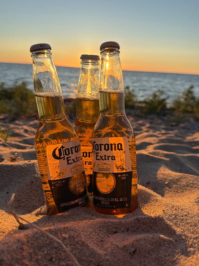 Three glass bottles of Corona Extra beer stand upright in the sand on a beach at Meremõisa, Estonia, at sunset. The bottles are partially filled with beer and illuminated by the golden light of the setting sun. The background consists of the sea, with a gentle horizon line, and a pale sky transitioning from yellow to blue. Some sparse green vegetation is visible along the shoreline. The sand is light brown and shows the texture of grains and small ripples.