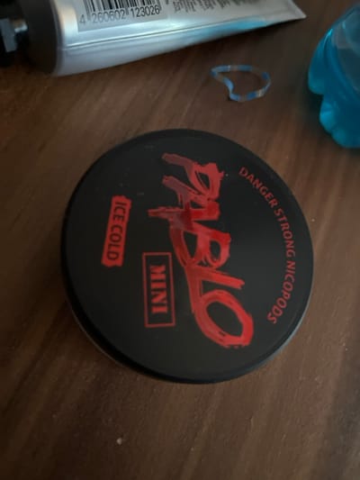 The scene depicts a collection of objects resting on a brown wooden surface with visible grain. In the foreground and central part of the image, a circular black container with a lid is prominently displayed. The lid features red lettering: "PABLO" in a large, textured font, "MINI" in a rectangular outline, "ICE COLD" in another rectangular outline, and "DANGER STRONG NICOPODS" curved along the upper right edge.
To the upper left of the container, a portion of a silver or light gray tube is visible, displaying a black barcode with the number 4260602123026. To the upper right, a segment of a light blue plastic object, likely a bottle or container, is present. Between the tube and the blue object, an irregularly shaped, translucent item with light blue and white stripes, resembling a plastic or rubber band, is visible.
No people, actions, or direct interactions are visible. The environment appears to be an indoor setting, possibly a table or desk. The specific location in Bad Nauheim, Germany, cannot be determined from the visual information provided within the image itself.