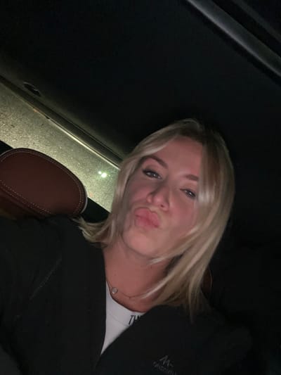 A blonde woman poses in a car, lips pursed, looking directly at the camera. She wears a white t-shirt and a black jacket. The interior of the car is visible, with the black roof and a brown leather seat with white stitching. The lighting is dim, creating shadows on her face and in the car. A window with blurry light is visible in the background.