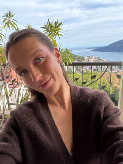A portrait photograph shows a woman with light skin, brown hair, and green eyes. She is wearing a brown cardigan and a delicate silver necklace. The background reveals a view of the city of Trebesin, Montenegro, with a coastline, buildings, and green vegetation visible through a metal railing with decorative leaf details. A plant with long, green leaves is in the foreground. The sky is partly cloudy.