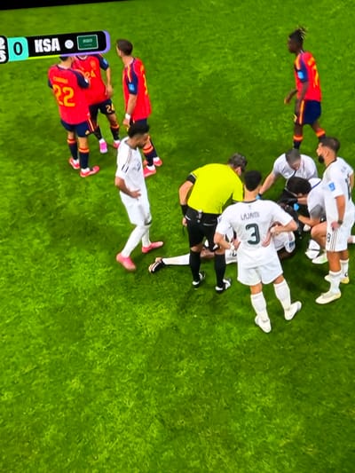 The image shows a soccer game on a green field. Several players in red and blue uniforms are gathered around a player on the ground. A referee in a yellow shirt is also present. There is a scoreboard in the background displaying "0 KSA". The scene appears to be an injury timeout during the match. The image does not provide any information about the city of Bykovo, Russia.